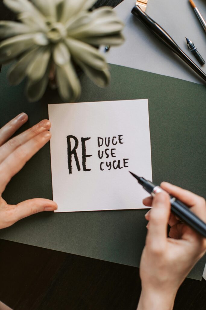 Close-up of hands writing a motivational 'Reduce Reuse Recycle' note on paper with a pen, surrounded by office items.